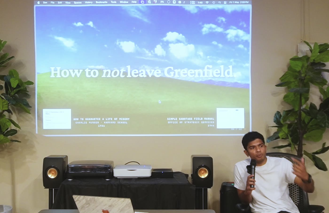 Wide shot from the talk. I'm seated with a mic, mid-gesture, the Windows-XP-green title slide - "How to not leave Greenfield." - projected behind me.