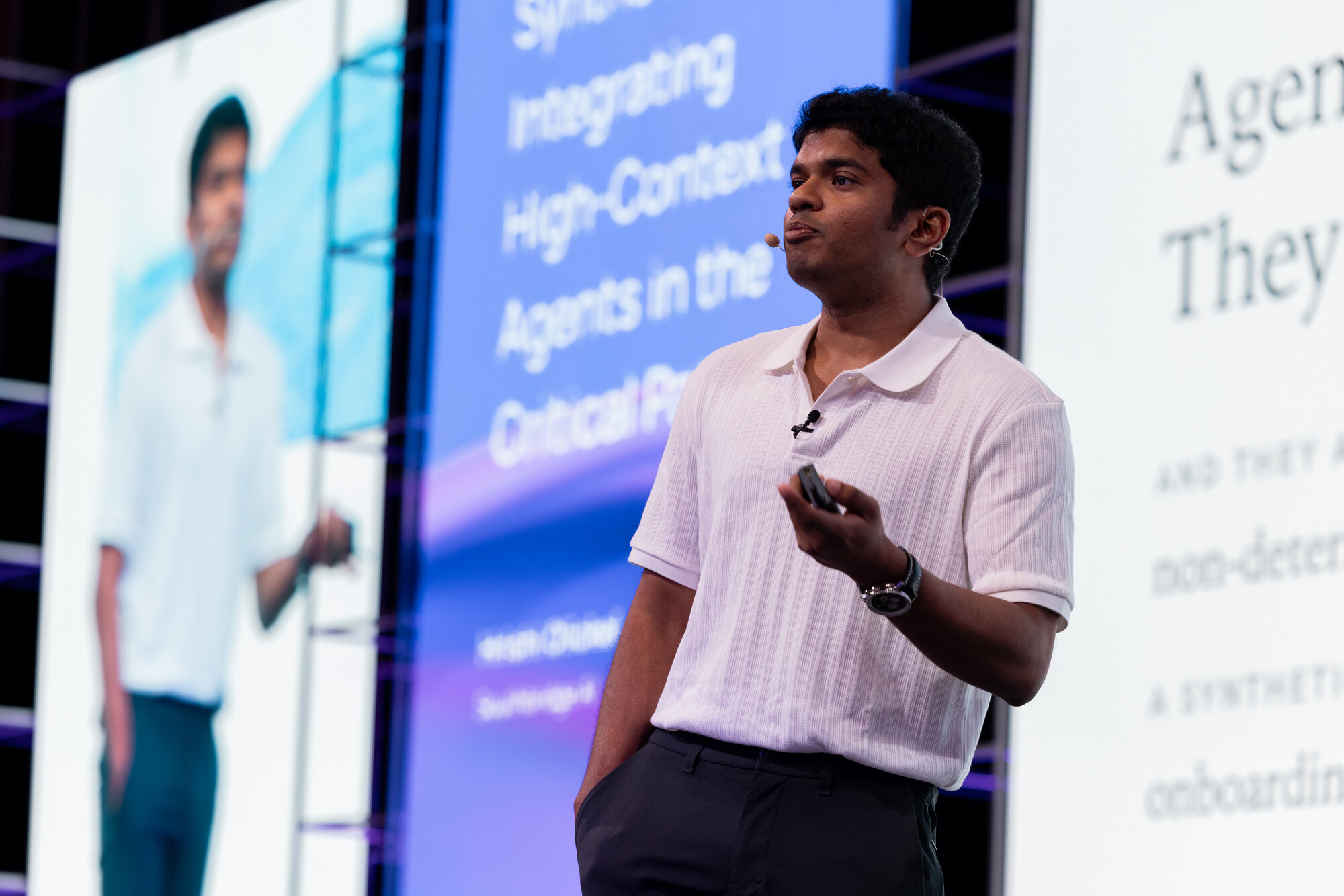 Mid-talk photo. I'm on stage in front of a large slide that reads "Agents are hires…" while a smaller monitor in front shows the title slide of the deck. I'm gesturing with the clicker.